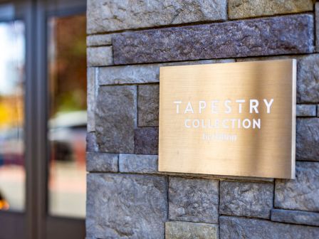 A stone-clad wall with a brass sign reading "TAPESTRY COLLECTION" near a glass door, suggesting a showroom or boutique boutique storefront.