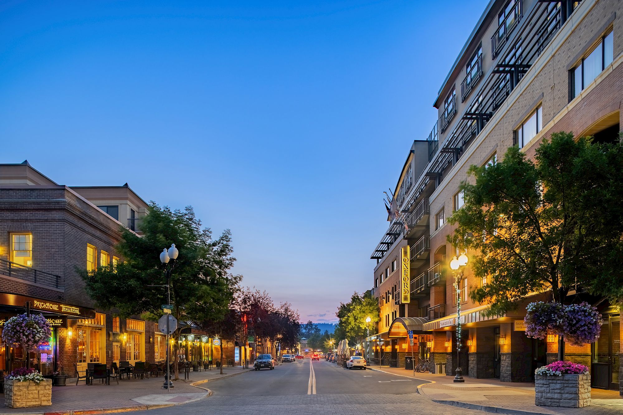 A pleasant street scene at dusk with shops and trees lining both sides, warm storefront lights glowing, and a clear blue sky above.