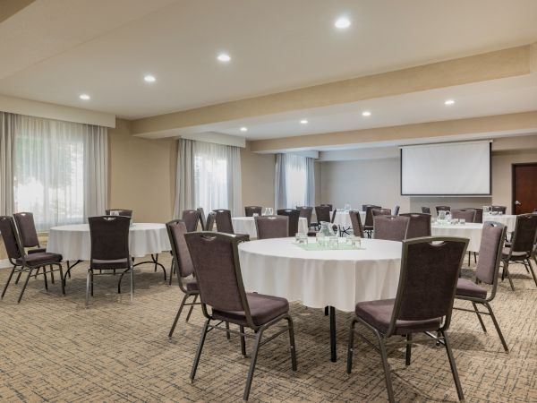 A conference room set up with multiple round tables, cushioned chairs, white tablecloths, and a projector screen at the front.