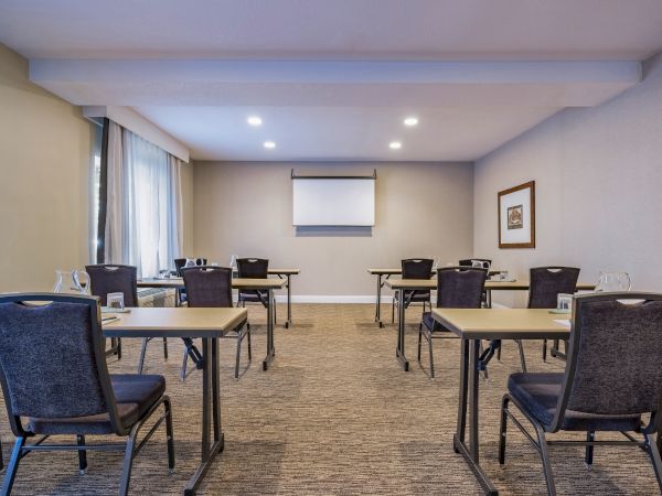 A cozy conference room with tables and chairs arranged for a meeting, a projector screen at the front, and a framed picture on the side wall.