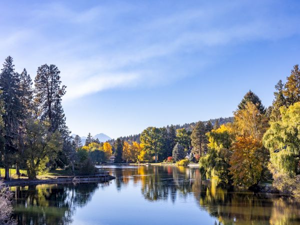 A serene lake framed by colorful autumn trees, clear blue sky, calm water mirroring the landscape, and distant hills glowing with fall hues.
