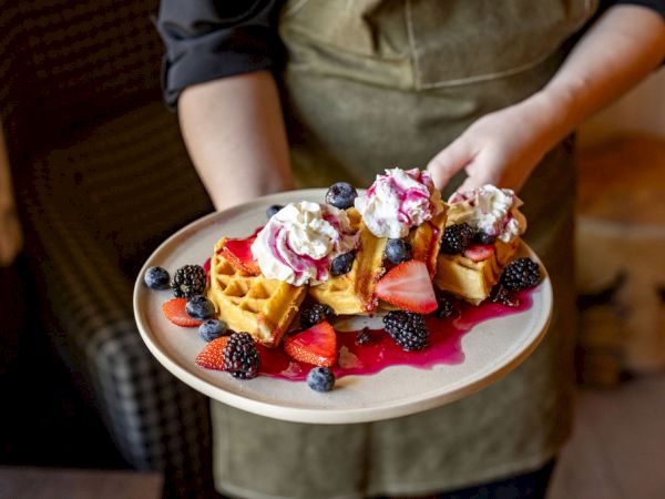 A plate of waffles topped with whipped cream and berries, drizzled with fruit sauces, being served by a waiter.