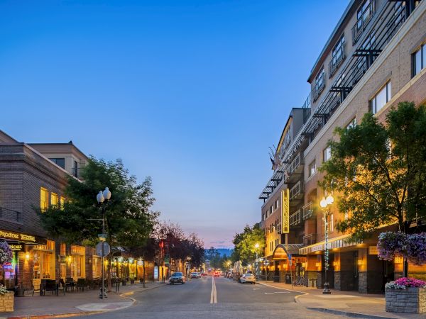 A calm street lined with shops and trees, warm lights glow from storefronts as evening settles over a pedestrian-friendly town.