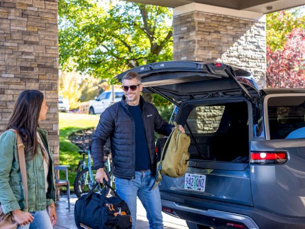A man and a woman loading bags into a blue SUV under a stone-covered canopy, preparing for a trip on a sunny day.