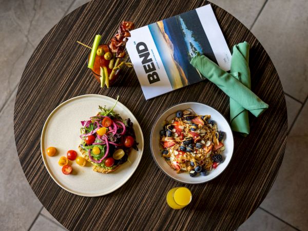 A round table with two plated meals, colorful salad and pasta, a yellow drink, a folded mint-green napkin, a Bend card, and a small toy plane on a tile floor.