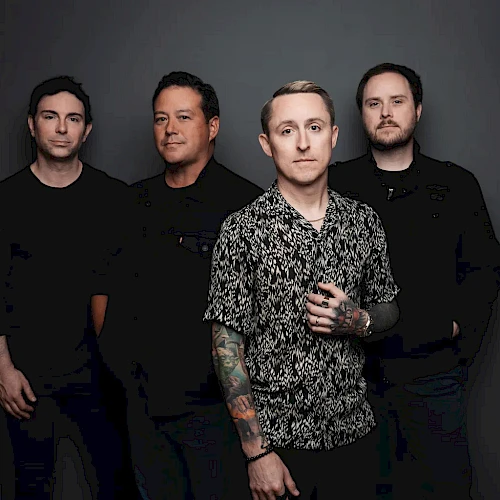 A four-member rock band posing for a photo, with one in a patterned shirt front and center, against a dark backdrop.
