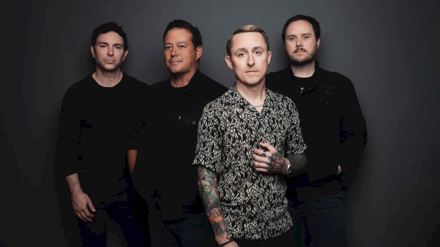 A four-member rock band posing for a photo, with one in a patterned shirt front and center, against a dark backdrop.