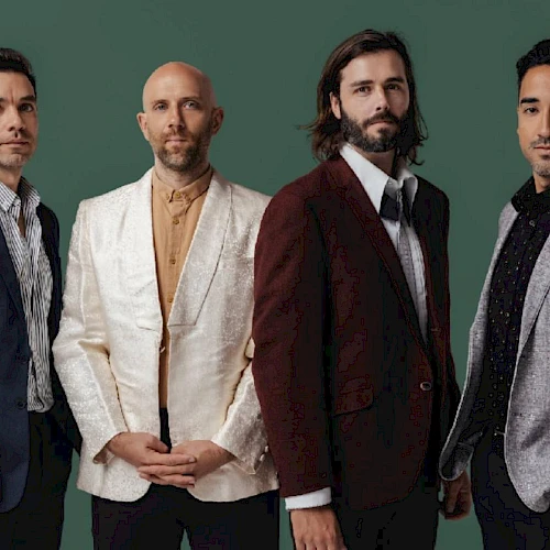 A four-member manly group posing in stylish suits against a green backdrop.
