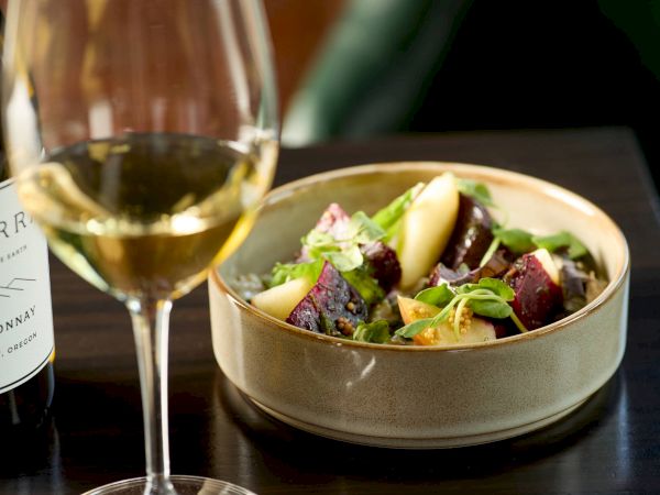 A glass of white wine beside a bowl of colorful salad with greens, beets, apples, and citrus, on a dark table. ending with a period.