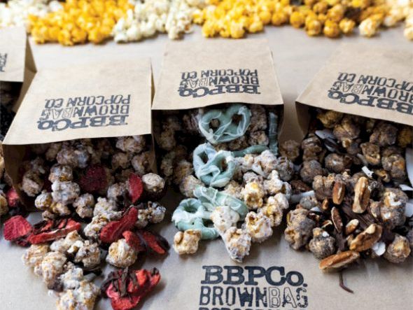 Assorted popcorn clusters in flavors, with colorful coatings and bags labeled &ldquo;BBPO Brown Bag Popcorn Co.&rdquo; for a tasty, decorative display.