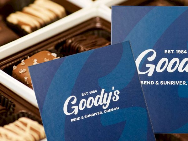 A box of assorted chocolates with a pair of Goody&rsquo;s business cards on top, featuring blue branding from Goody&rsquo;s in Bend, Oregon.