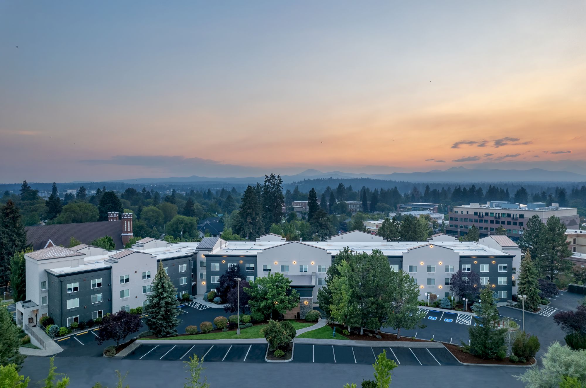 An aerial view of a multi-building apartment complex with parking lots, trees, and a sunset sky over the treeline.