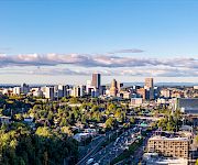 A wide city skyline with tall office towers, tree-filled streets, cars on highways, and a bright blue sky with clouds.