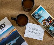 On a wooden table, two coffee cups and travel brochures for the Adirondacks and Newberry Hotel are spread out.