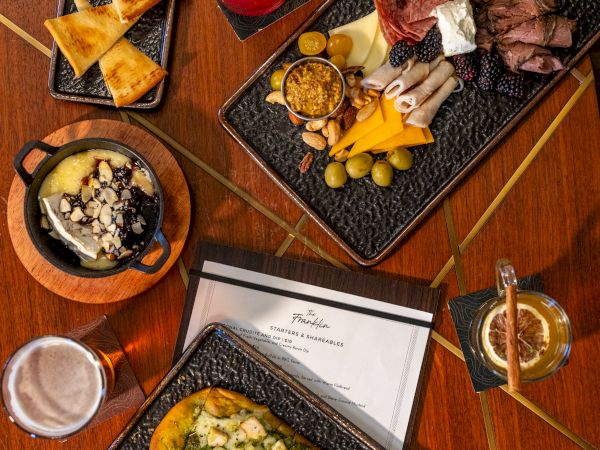 A stylish spread of nachos, a vibrant veggie curry, roasted veggies and dip, a hearty fish dish, fresh fruit slices, and a bright red cocktail, all on a wooden table.