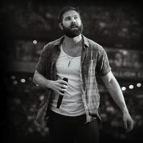 A bearded man on a stage holds a microphone while speaking or singing in front of a blurred audience, likely at a concert.
