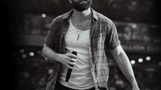 A bearded man on a stage holds a microphone while speaking or singing in front of a blurred audience, likely at a concert.