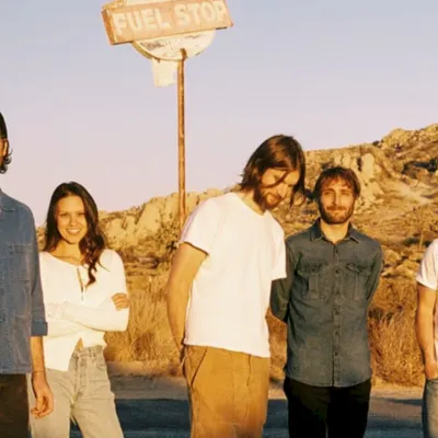 A group of five friends stands in a desert landscape by a &ldquo;FUEL STOP&rdquo; sign at sunset.