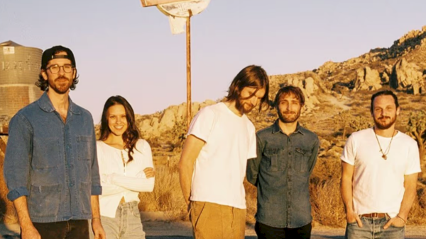 A group of five friends stands in a desert landscape by a &ldquo;FUEL STOP&rdquo; sign at sunset.