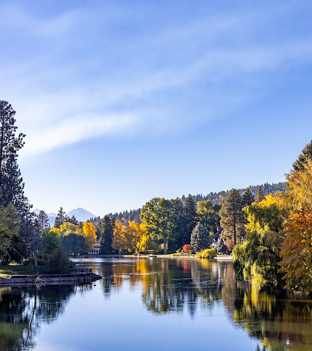 A calm lake with reflections, surrounded by autumn trees and distant hills under a blue sky with wispy clouds.
