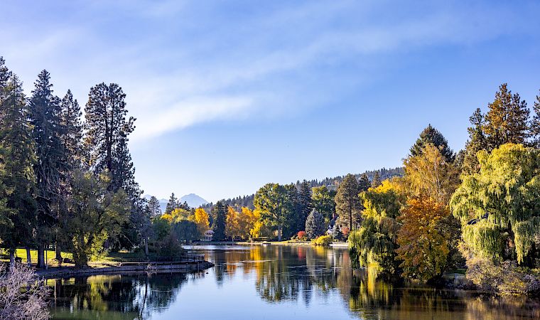 A calm lake with reflections, surrounded by autumn trees and distant hills under a blue sky with wispy clouds.