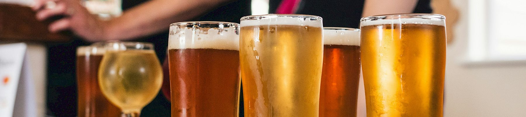 Several beers in clear glasses on a wooden board while people in the background pour and cheer in a brewery taproom.