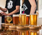 Several beers in clear glasses on a wooden board while people in the background pour and cheer in a brewery taproom.