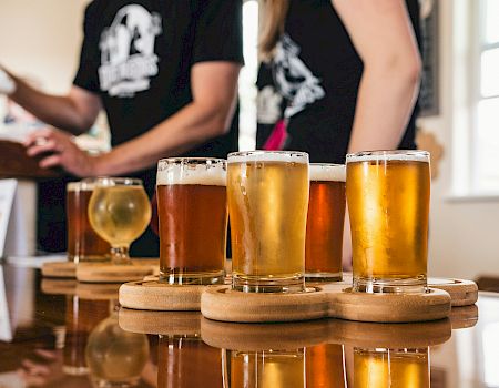 Several beers in clear glasses on a wooden board while people in the background pour and cheer in a brewery taproom.