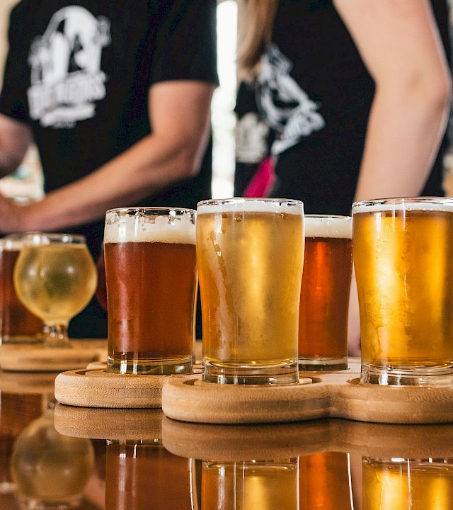 Several beers in clear glasses on a wooden board while people in the background pour and cheer in a brewery taproom.