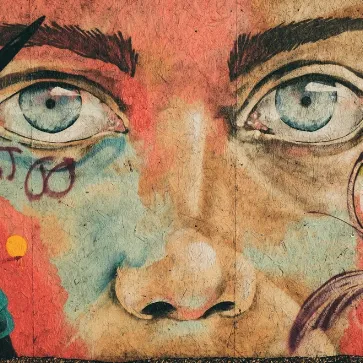 A close-up of a stylized, graffiti-style face with intense blue eyes and mixed red, yellow, and teal paint.