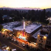 Aerial dusk view of a small town with lit streets, a glowing central building, parked cars, and mountains in the background.