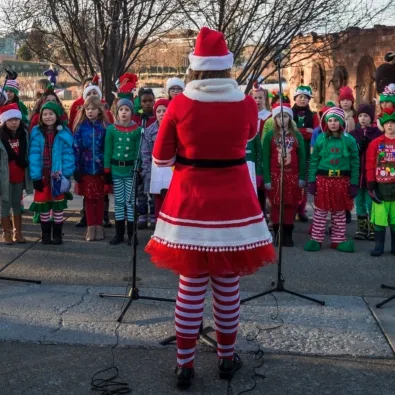 A woman in a Santa-style outfit leads a group of costumed kids singing outdoors in winter, with microphones set up.