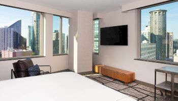 A modern hotel room with large windows showing a cityscape, a TV mounted on the wall, and a bed and chair.