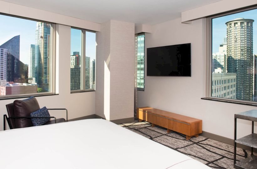 A modern hotel room with large windows showing a cityscape, a TV mounted on the wall, and a bed and chair.