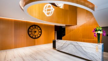 A modern, elegant lobby with a marble front desk, wood paneling, a large clock on the wall, and a curved ceiling fixture.
