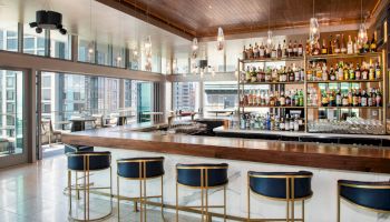 A modern bar with a sleek design, featuring a stocked bar shelf, high stools, and large windows offering a city view.