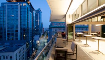 A modern outdoor balcony on a high-rise, overlooking city lights at dusk, with seating and open glass doors to the interior space.