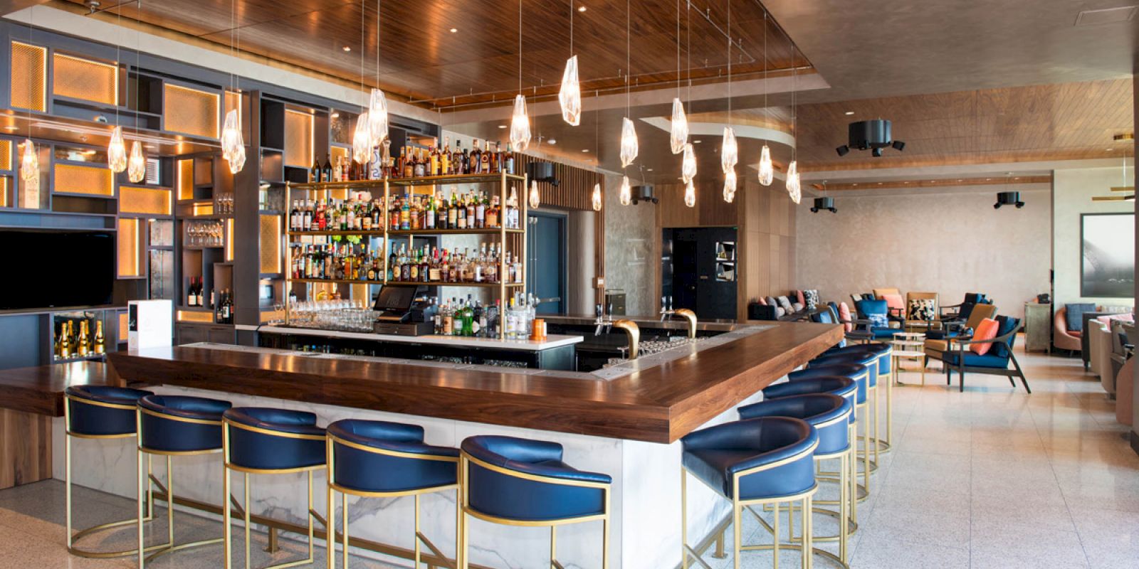 The image shows a modern bar with a sleek counter, blue stools, a well-stocked liquor display, pendant lighting, and a lounge area.