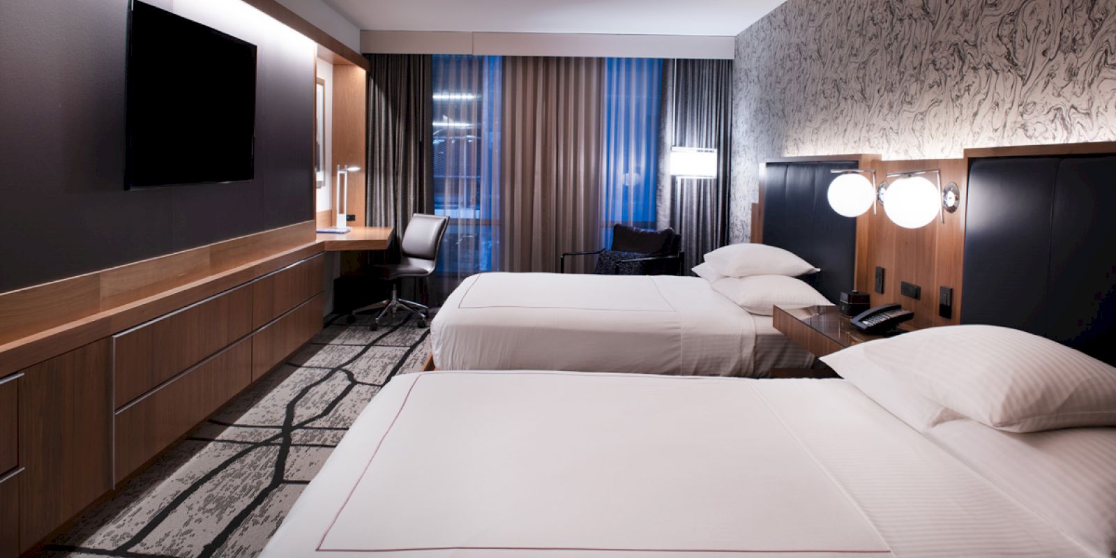 A modern hotel room with two beds, a flat-screen TV, desk area, and patterned wallpaper.