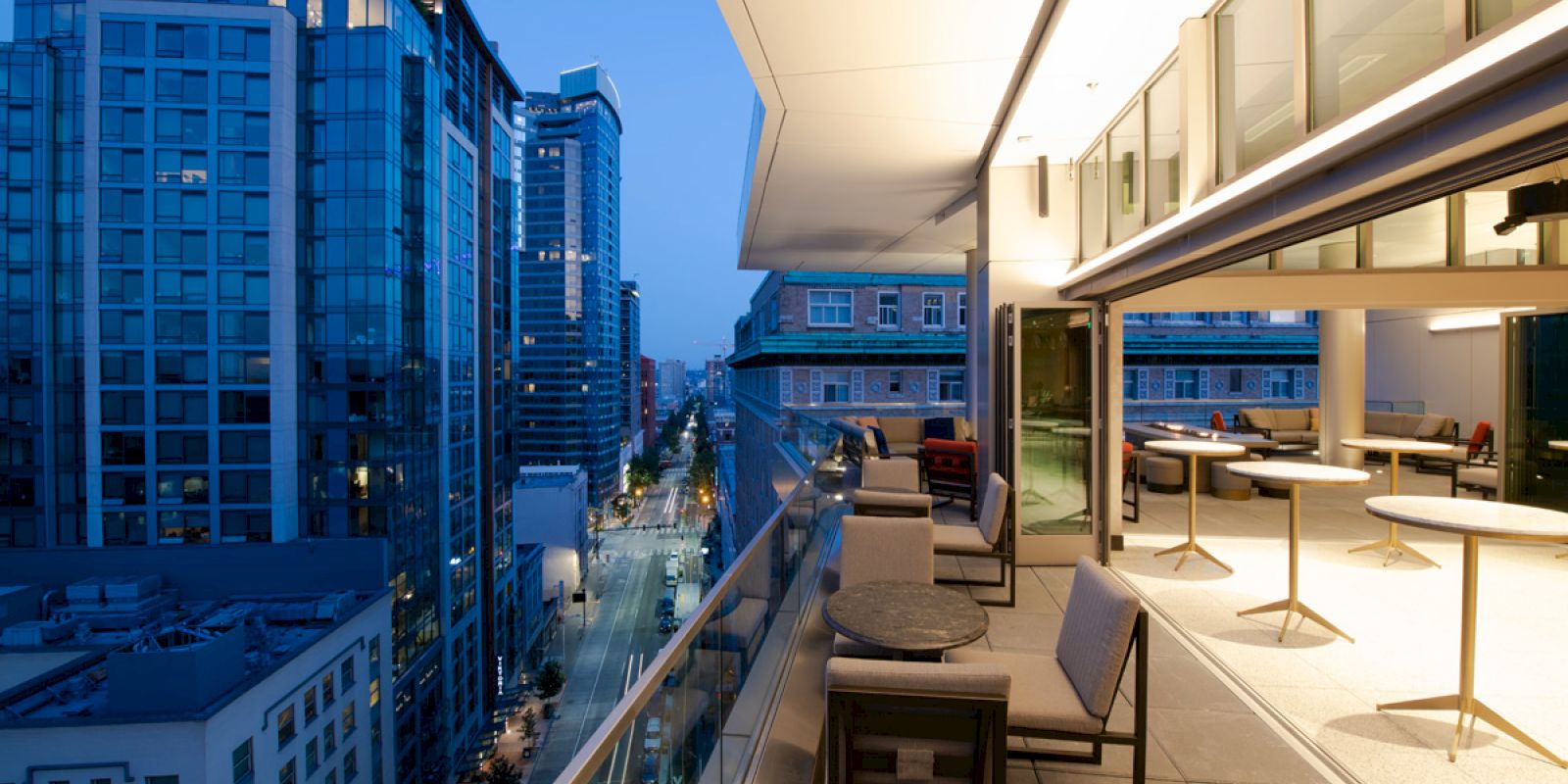 A modern urban balcony with seating overlooks a city street lined with tall buildings, illuminated in a twilight setting.