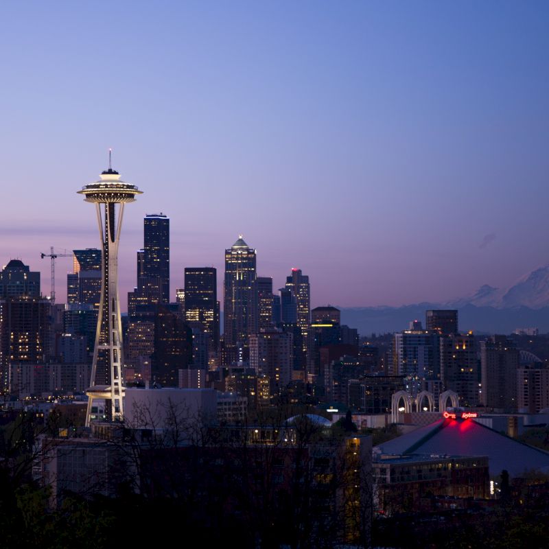 Seattle skyline at dusk featuring the Space Needle with Mount Rainier in the background and city lights beginning to glow.