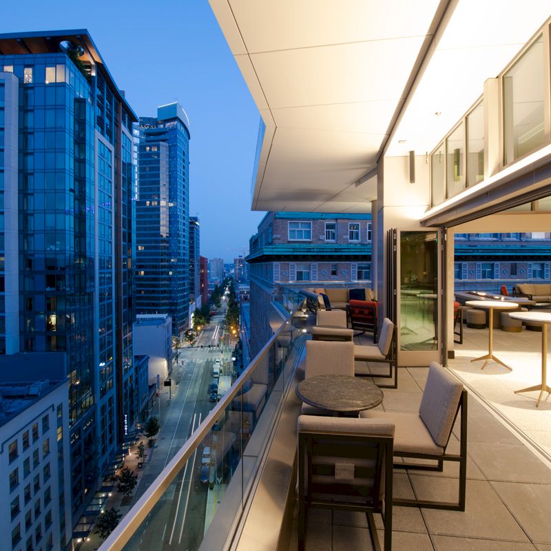 A modern rooftop terrace with seating overlooks a city street and high-rise buildings during dusk, illuminated by soft, ambient lighting.