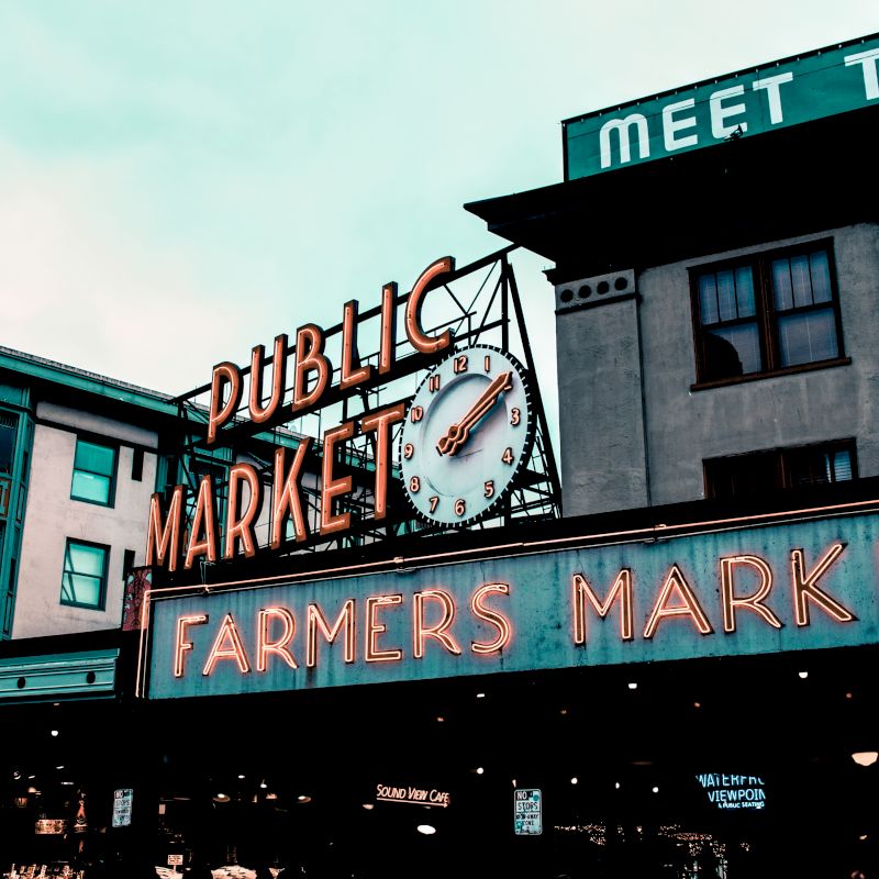 The image shows a neon sign reading "Public Market" with a clock, and "Farmers Market," against a building backdrop.