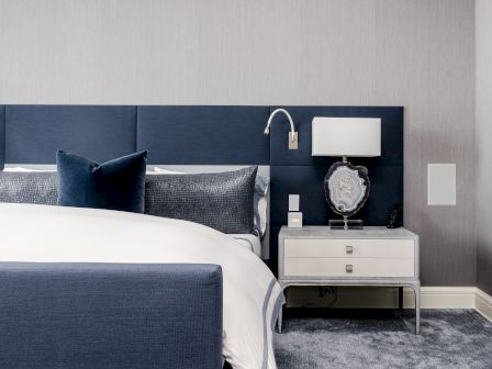 A modern bedroom features a blue headboard, a bed with white and blue bedding, and a nightstand with a lamp and decorative item.