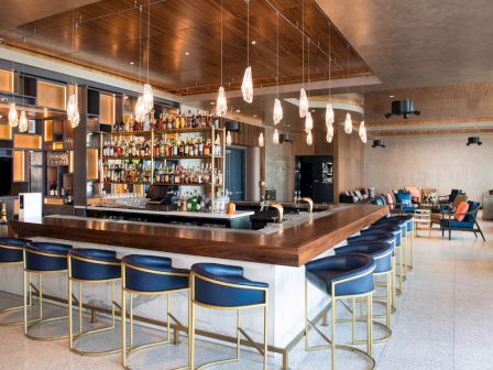A modern bar with a wooden counter, high blue chairs, bottle shelves, and hanging lights, with a lounge area in the background.