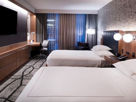 A modern hotel room with two beds, a wall-mounted TV, a desk with a chair, and stylish lighting and decor.