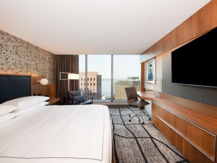 A modern hotel room with a large bed, desk, chair, TV, and city view through large windows, featuring a patterned accent wall.