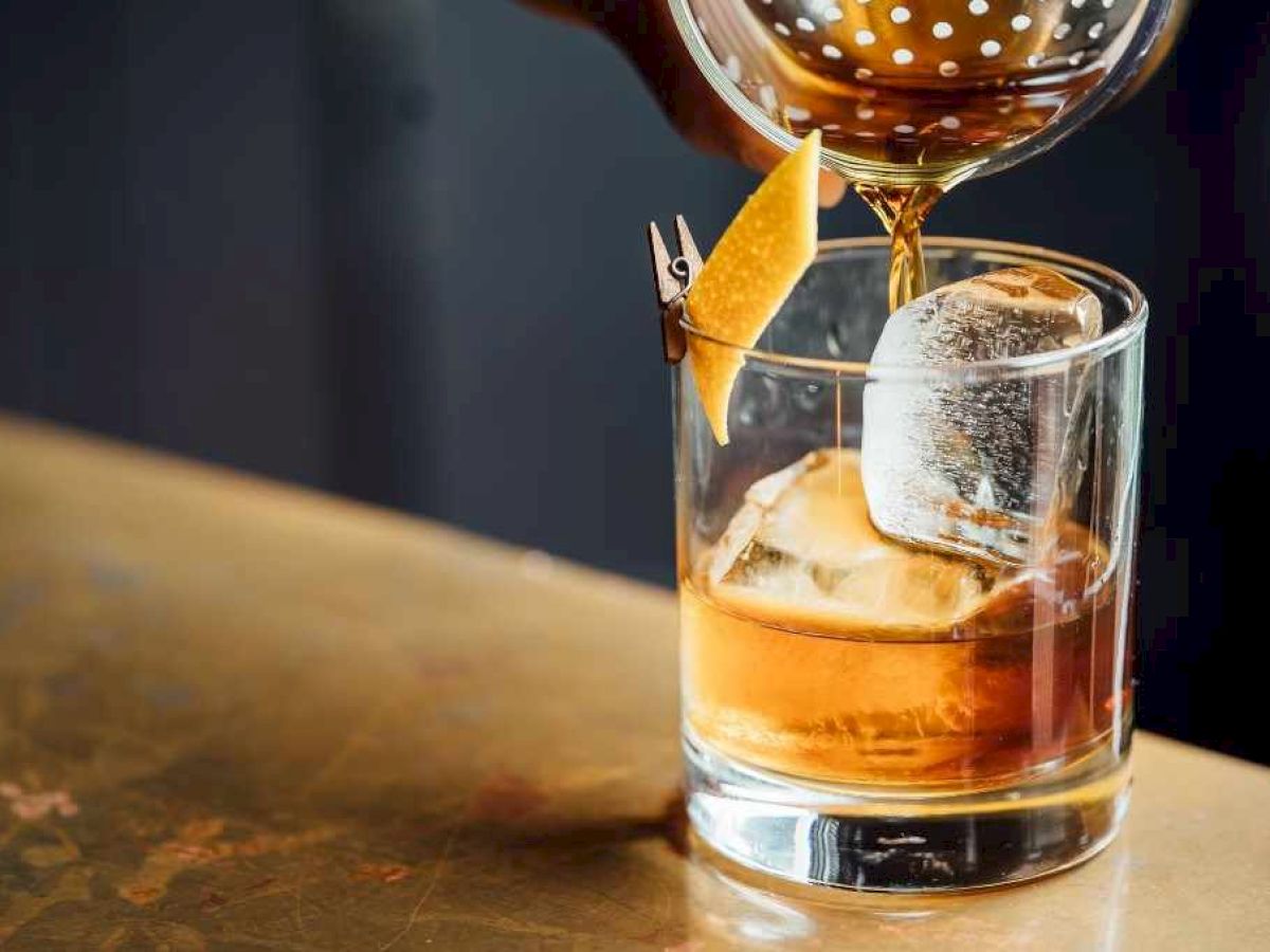 A cocktail is being poured into a glass with ice and a lemon twist on a bar.