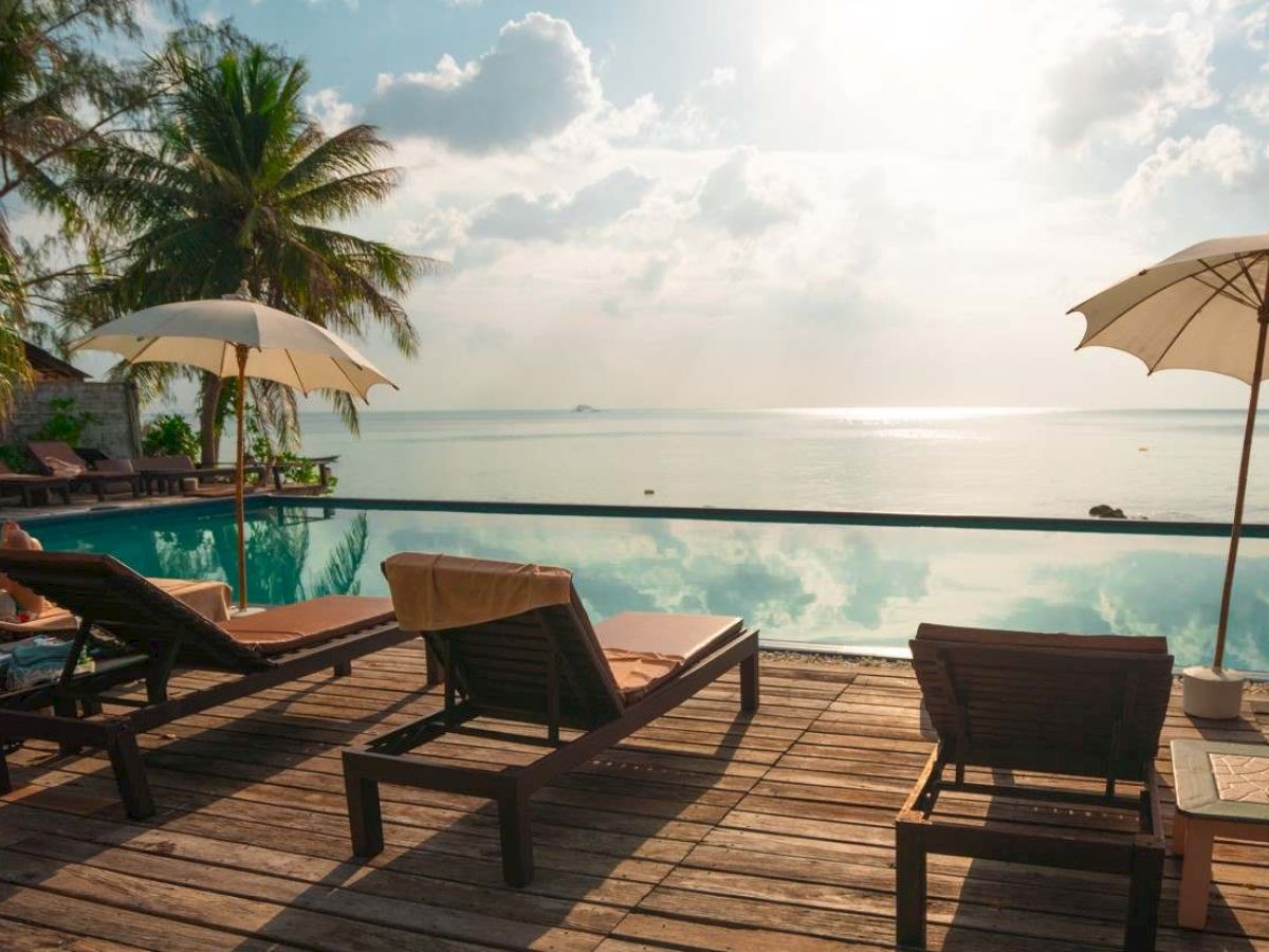 Lounge chairs and umbrellas by a poolside with a view of the ocean, palm trees, and a serene sky in the background.