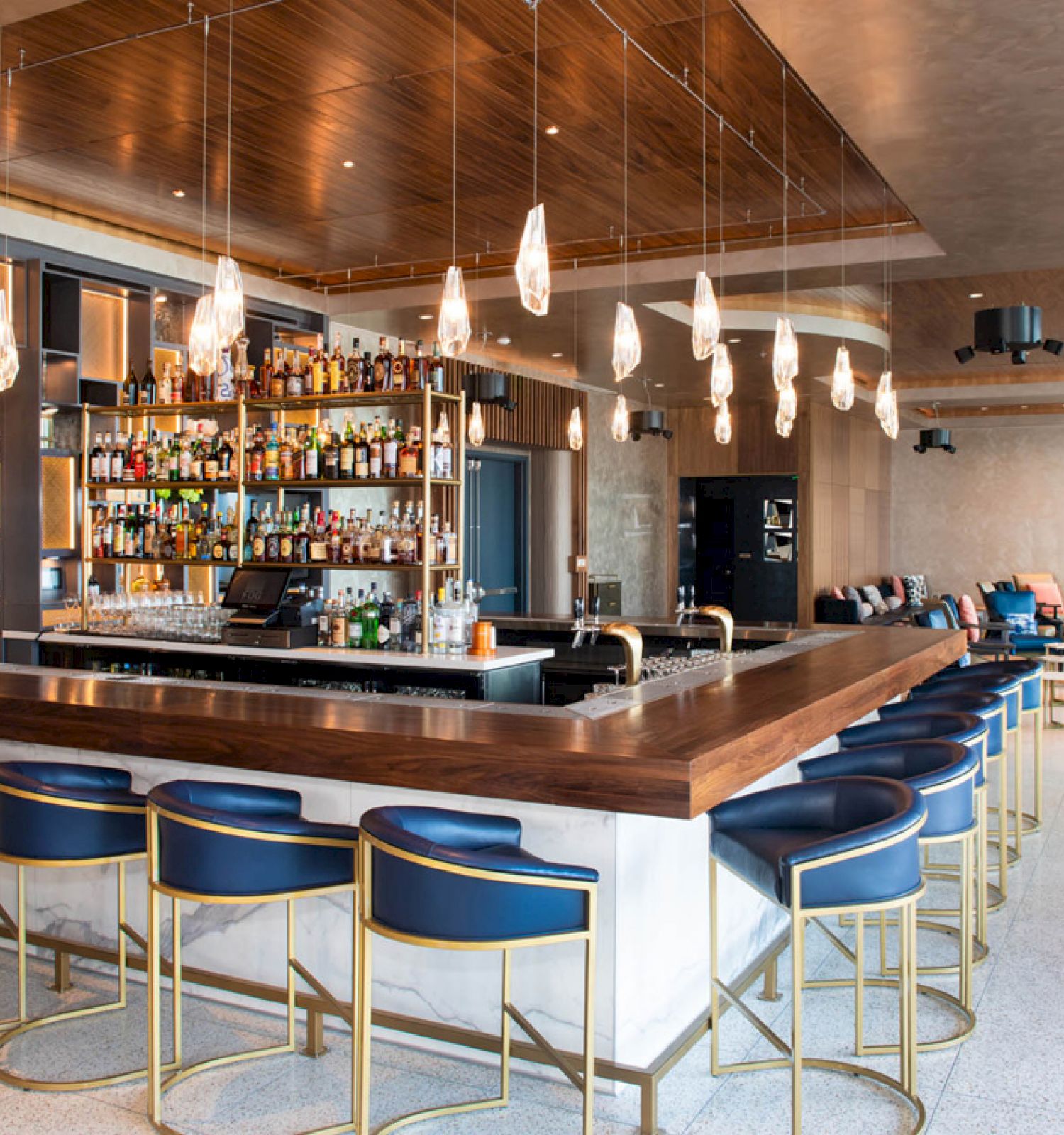This image shows a modern bar with a wide selection of bottles, stylish seating, pendant lights, and a cozy seating area in the background.